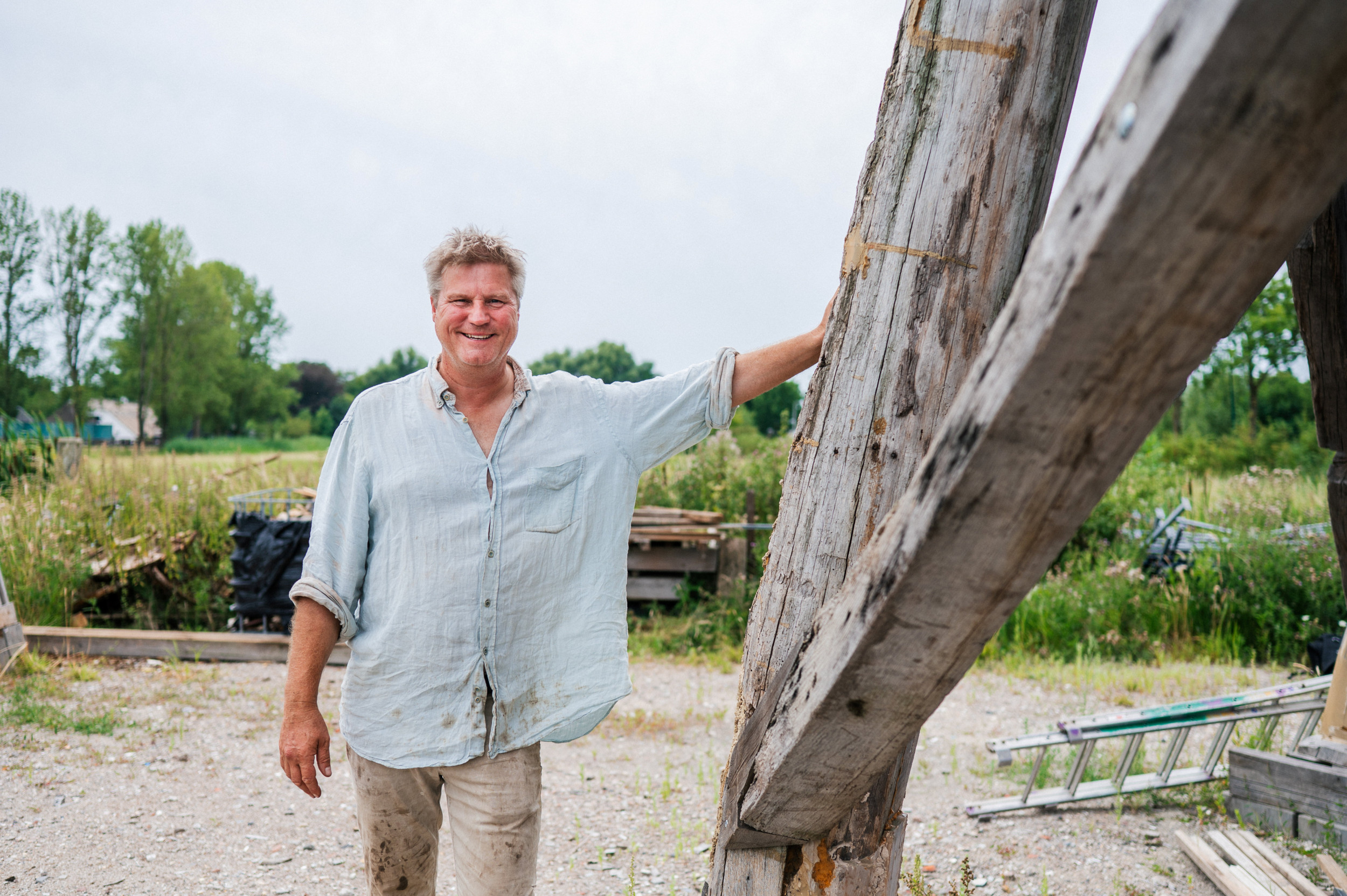Man in kluskleding bij dikke houten balken, in een groene omgeving.