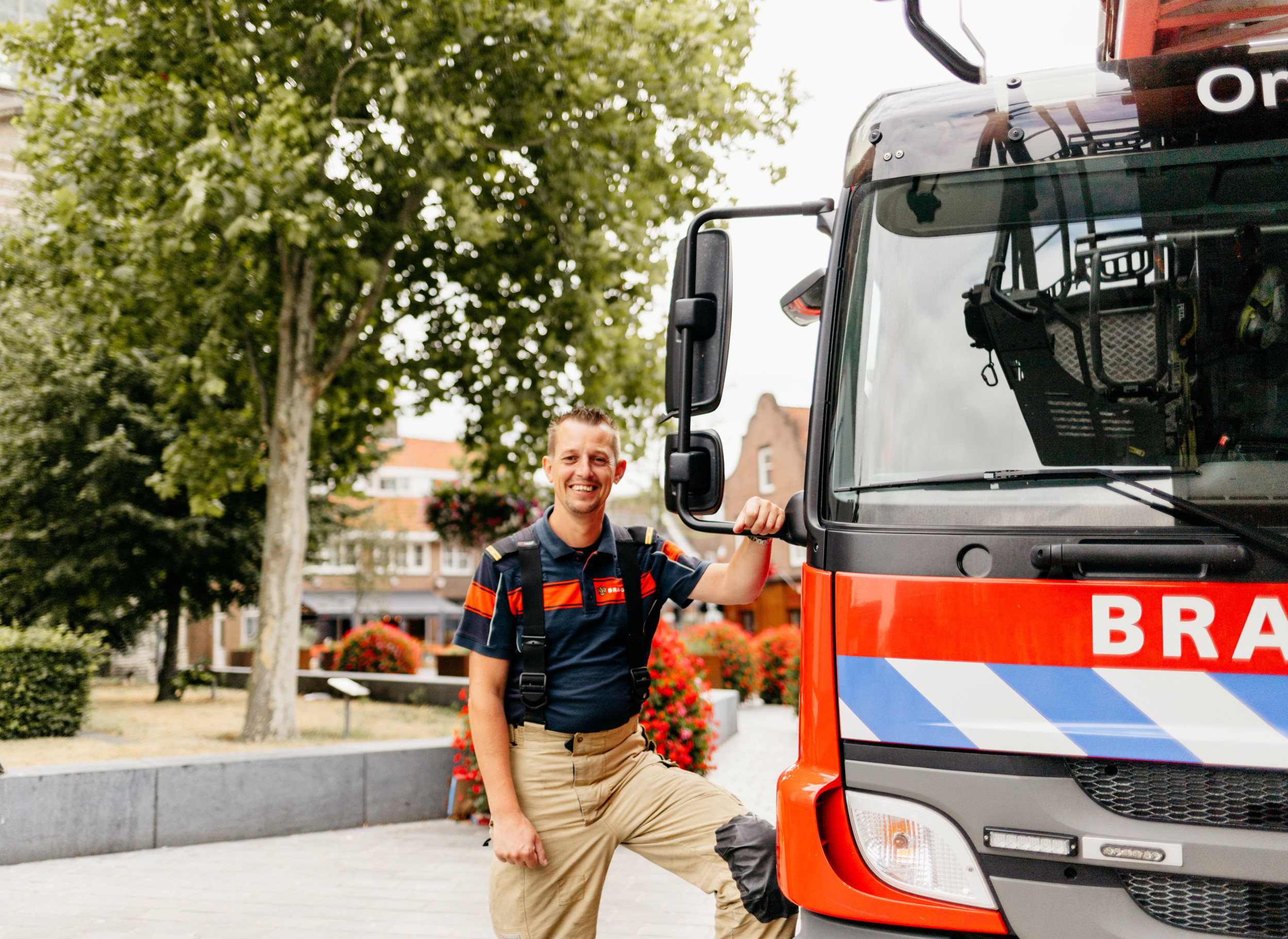 Brandweerman naast een brandweerwagen. Op de achtergrond bomen en huizen.