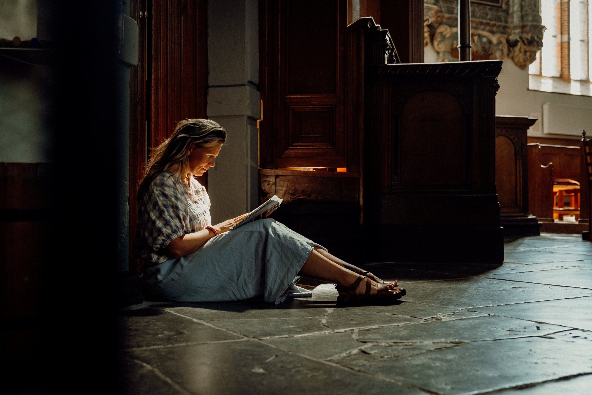 Vrouw met blond haar en een blauwe rok, lezend in een boek op de vloer van een oude kerk.
