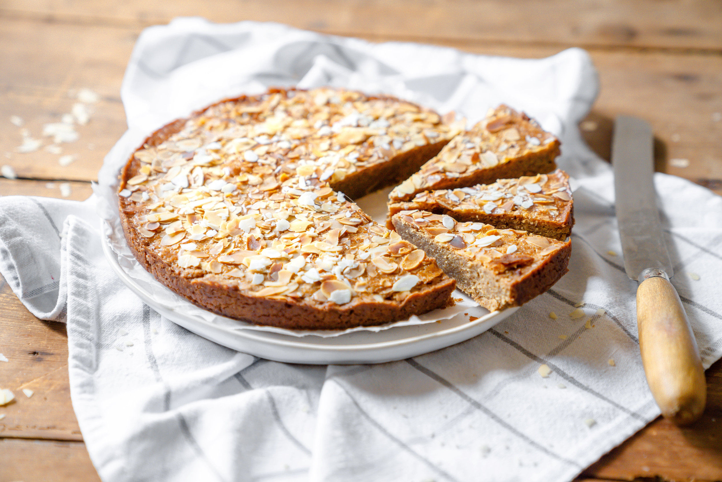 Wit bord op een wit-grijs geruite theedoek. Op het bord een platte, ronde en goudbruin gebakken koek met amandelschaafsel erop. 