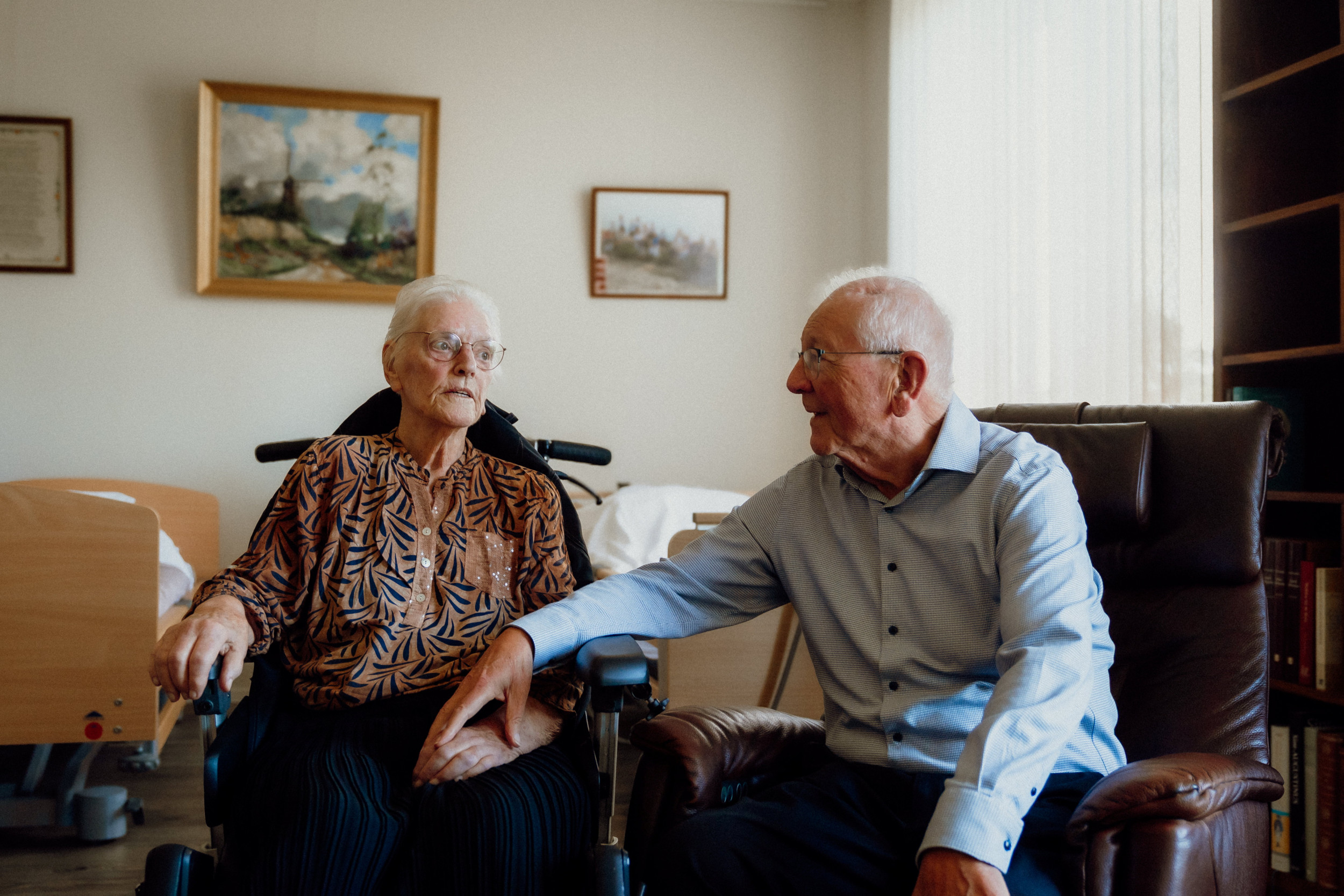 Oude vrouw in een rolstoel naast een oude man in een stoel, met een hand op haar hand.