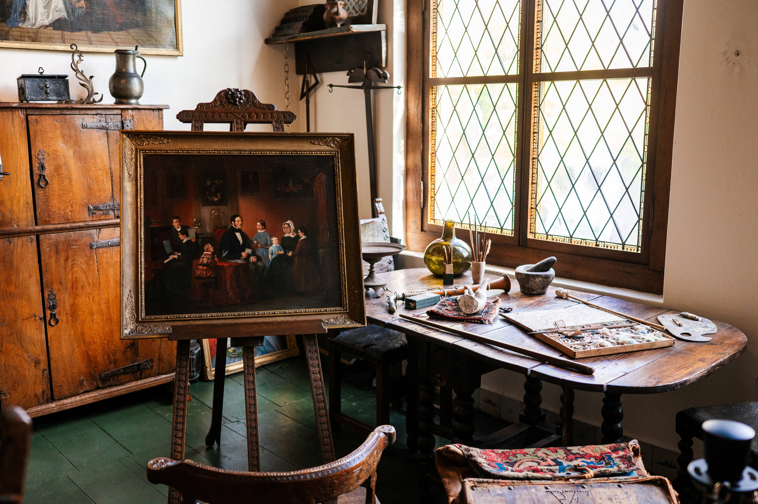 Antieke meubels, een ingelijst schilderij op een schildersezel en schildersbenodigdheden op een tafel voor een raam.