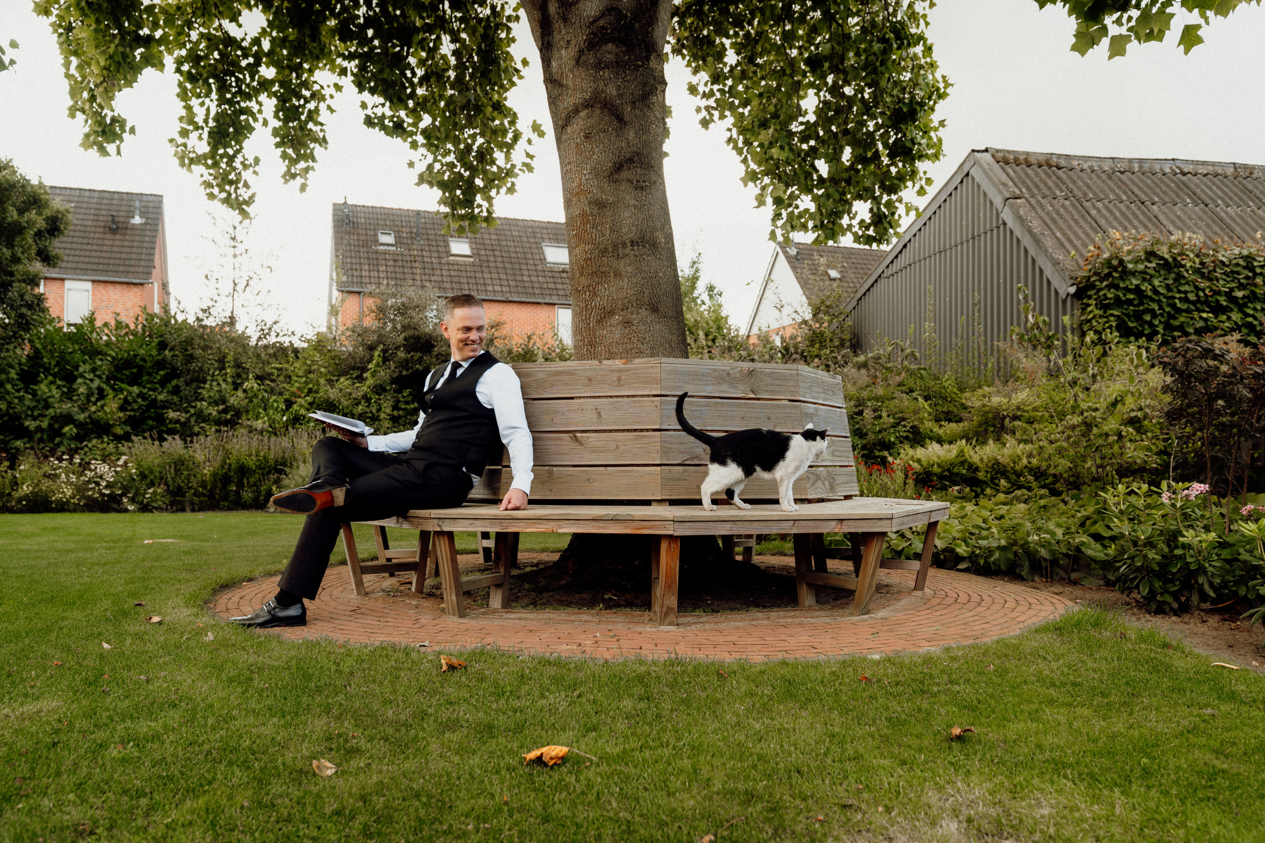 Man gekleed in zwarte pantalon, zwart gilet, zwarte das, wit overhemd en zwarte sokken en schoenen; zittend op een bankje onder een boom. Op het bankje loopt een zwart-witte kat.