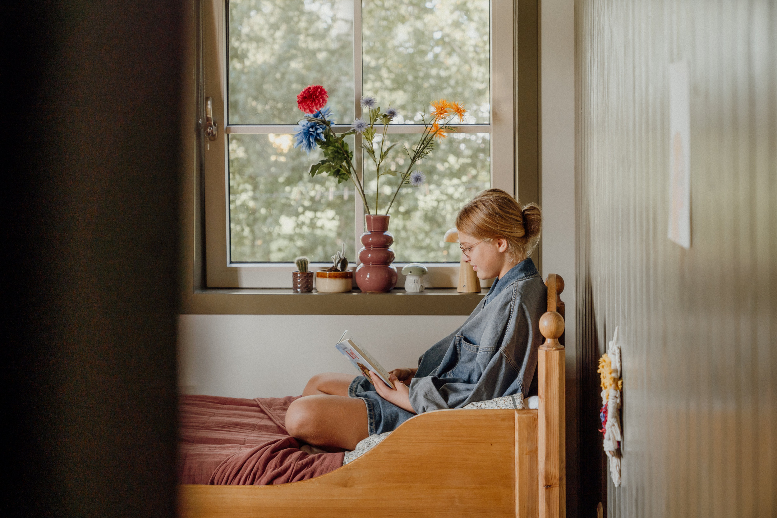 Een meisje zit op een bed, voor het raam, te lezen. De muren zijn oudgroen.