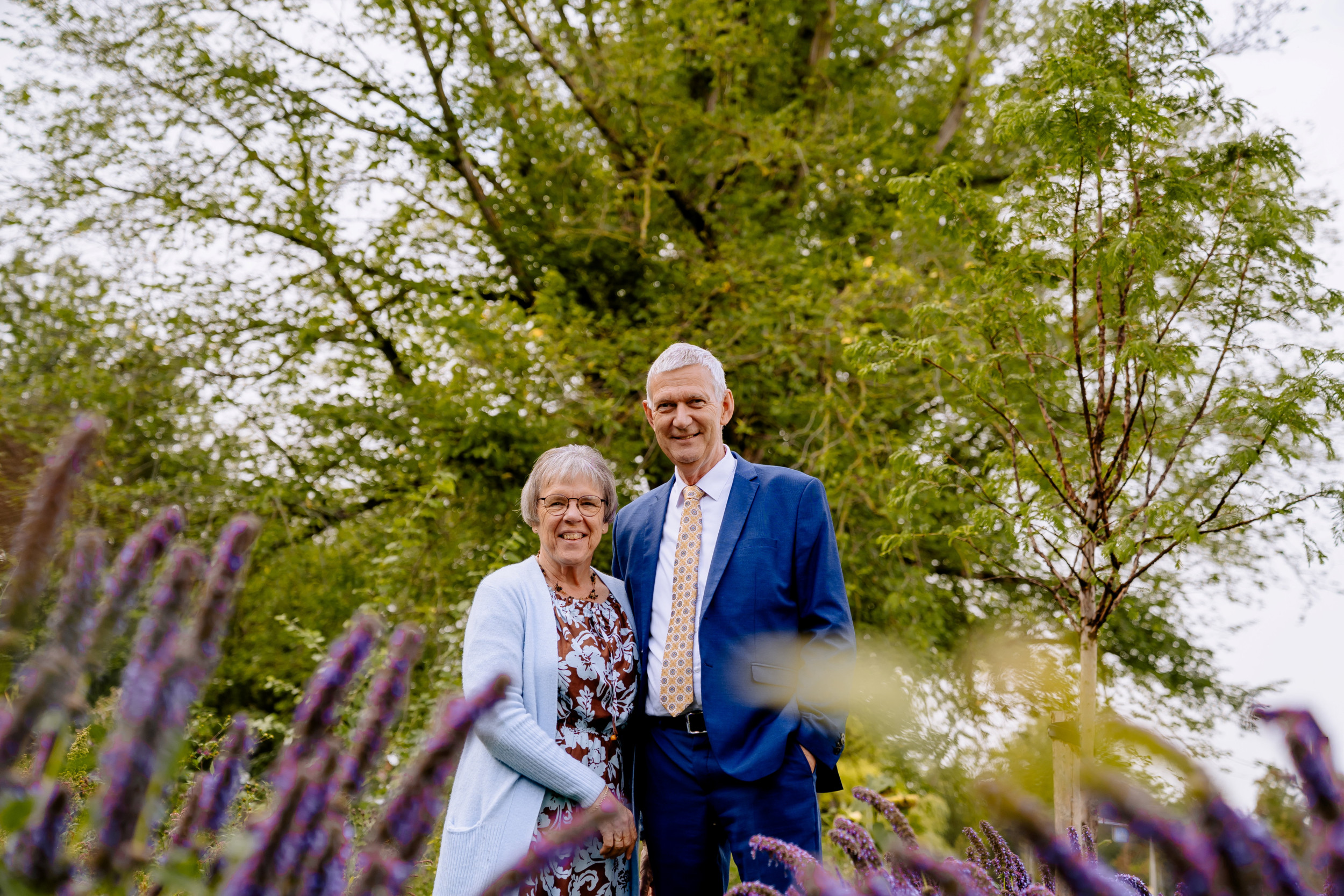 Vrouw met lichtblauw vest naast een man in een blauw kostuum in een tuin met bomen.